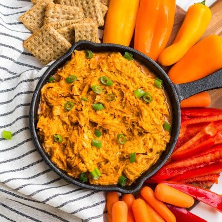 small cast iron skillet with cooked buffalo chicken dip. Dip has sliced green onions on top, and there are crackers and cut vegetables around the base of the skillet.