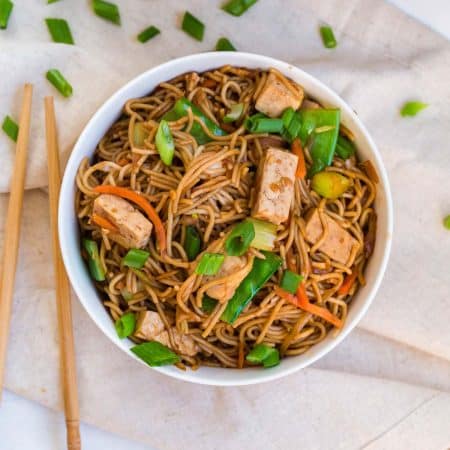 top down image of a white bowl filled with vegetable lo mein. You can see chunks of tofu, brown rice noodles, cut snow peas, and sliced green onions. In the background are chopsticks and more cut green onions on a white counter