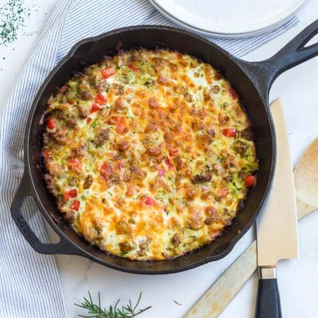 top down view of a cast iron skillet with ground turkey sausage, red peppers, zucchini, and browned cheese on top. Knife and wooden spoon are in the background.