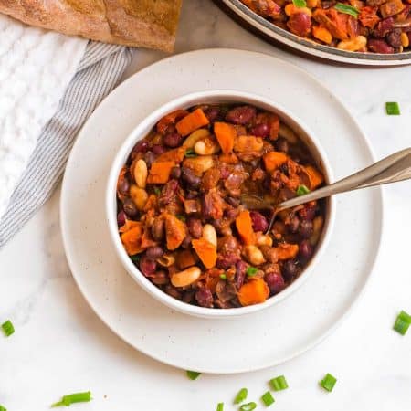 white plate with white bowl on top. The bowl has vegan chili inside, and you can see carrots, kidney beans, cannelini beans, and black beans alongside chunks of tomato.