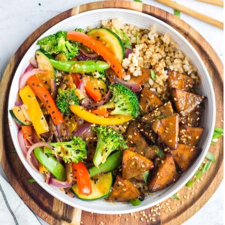 white bowl of cooked teriyaki tofu and vegetables with coconut rice. Bowl is on a wood platter with sesame seeds sprinkled around.