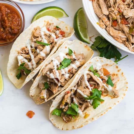 three carnitas tacos with shredded pork carnitas, tomatoes, and cilantro on top and garnished with swirls of sour cream. Off to the side are a clear dish of salsa and a large bowl of finished shredded carnitas