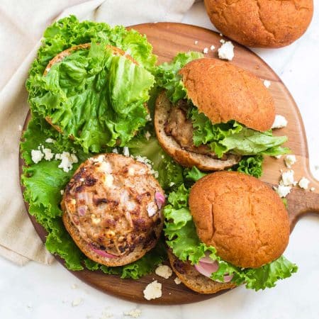 top down view of hand cut whole wheat burger buns with green pieces of lettuce. On the buns are cooked turkey burgers with onion and feta cheese. Burgers are placed on a wood platter