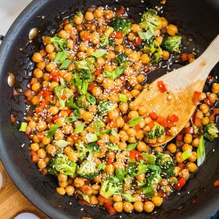 Top down view of cast iron skillet with garbanzo beans, red peppers, broccoli, sliced green onions, and sesame seeds cooking.