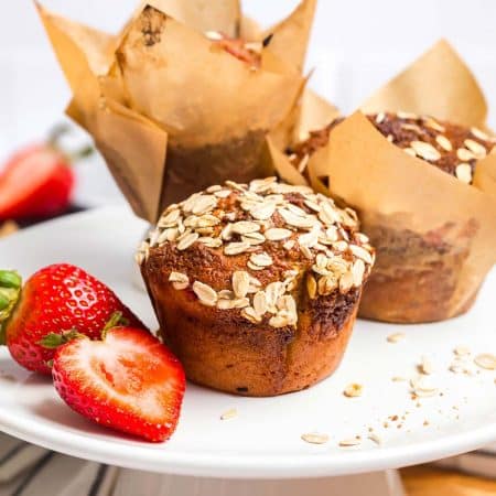 Cooked Strawberry muffin on a white cake plate, there are two more muffins in the background with baking paper around them, and 1 1/2 strawberries off to the left of the muffin.