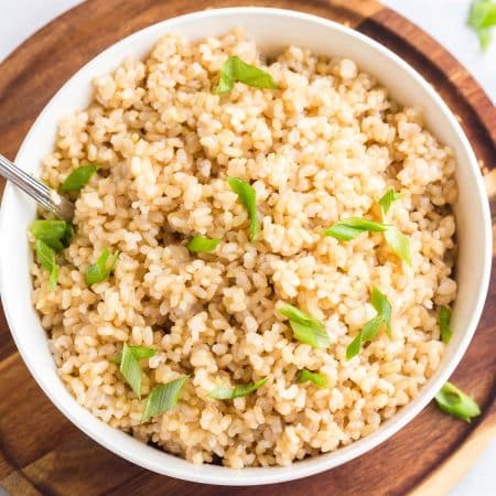 white bowl of cooked coconut brown rice with cut chives in bowl. Bowl is on a wood platter.