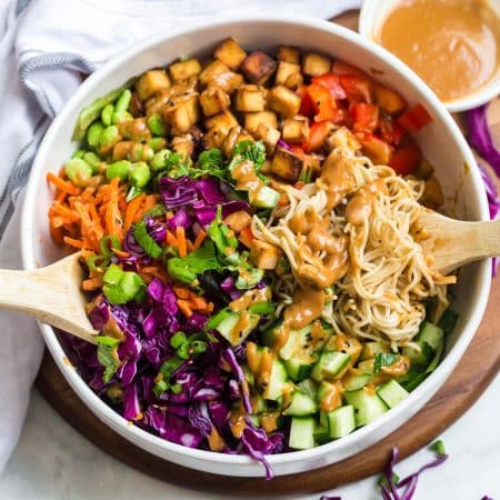 top down image of white bowl of Spring Roll Salad. Shredded carrots, chopped purple cabbage, diced cucumbers, cooked brown rice ramen, cubed air fried tofu, and edamame beans with a creamy peanut sauce on top.