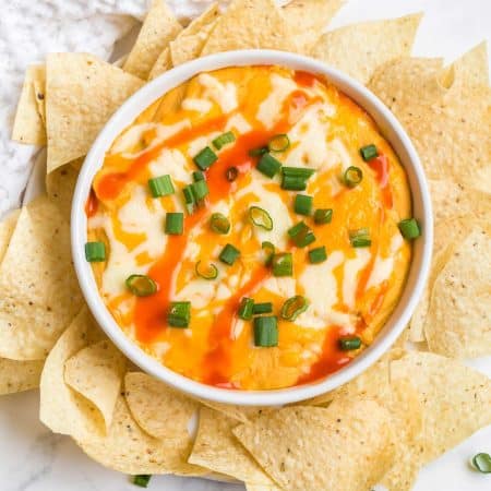 white bowl with buffalo cauliflower dip inside. Dip has swirls of buffalo sauce and chopped green onions on top. Around the bowl are white corn tortilla chips.