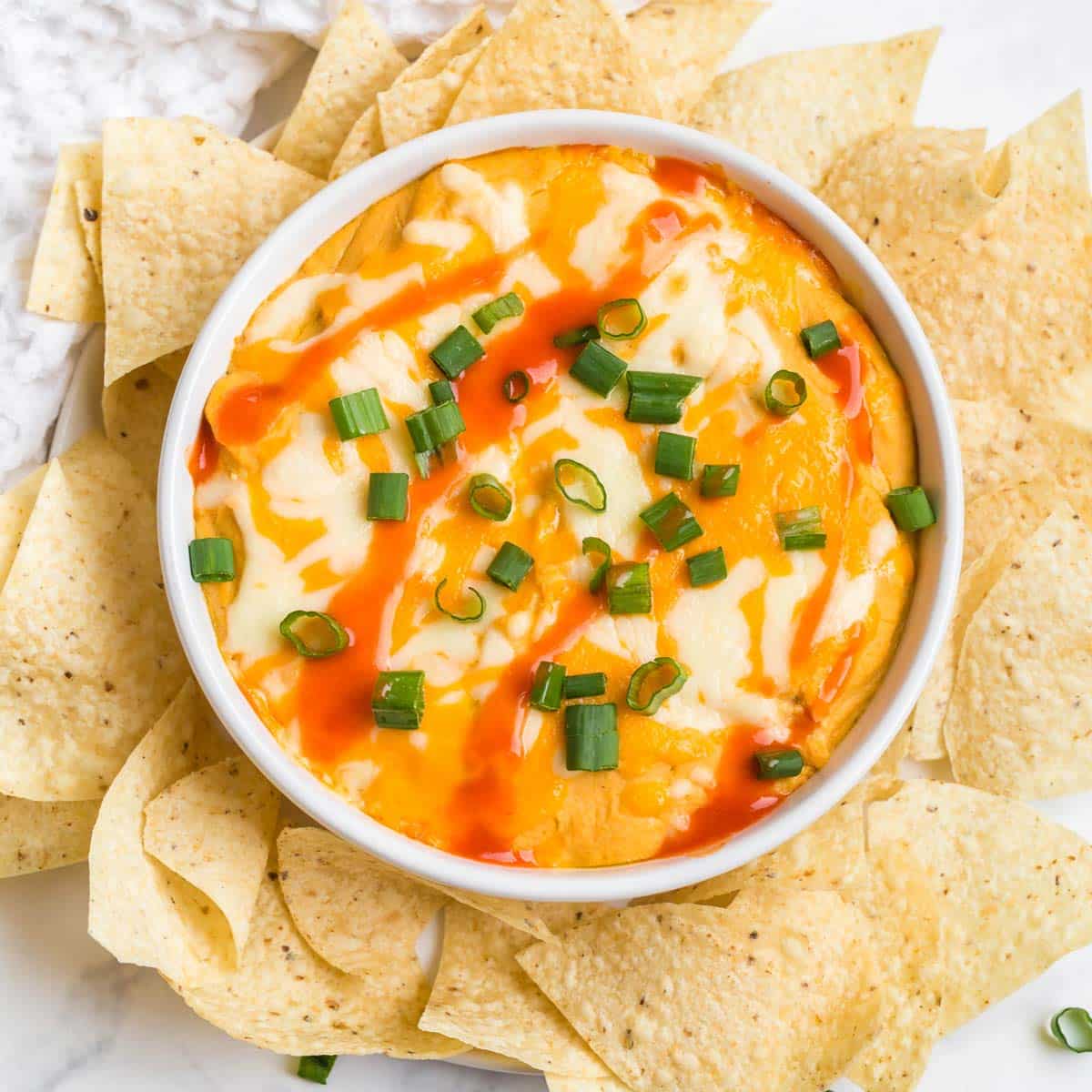 white bowl with buffalo cauliflower dip inside. Dip has swirls of buffalo sauce and chopped green onions on top. Around the bowl are white corn tortilla chips. 