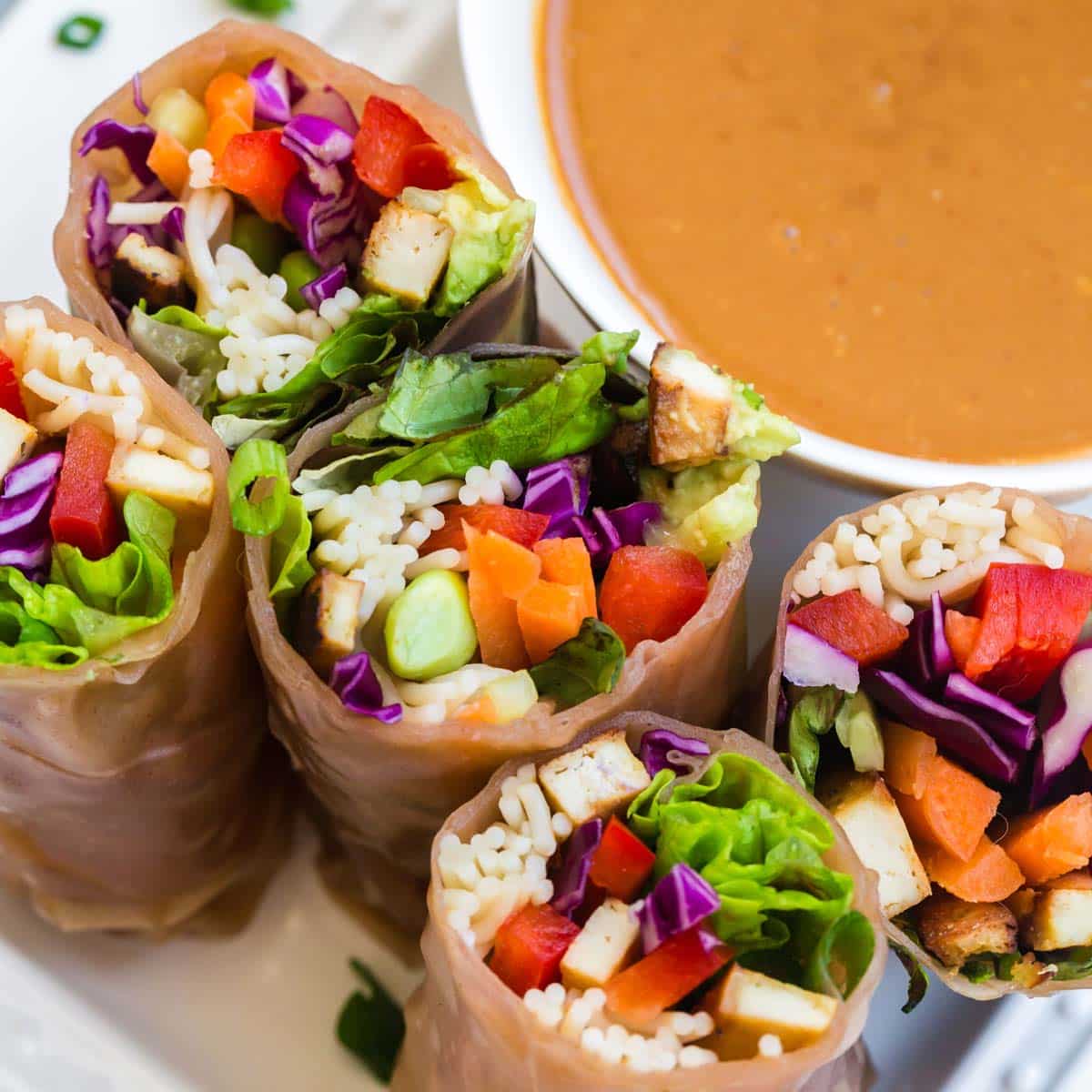 close up of vegetable spring rolls cut in half. The rolls have colorful vegetables in them and a small bowl of peanut sauce on the top right side.