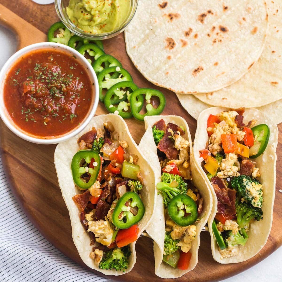 three breakfast tacos, a white dish of salsa, sliced jalapenos, a clear dish of guacamole, and tortillas arranged on a wood dish