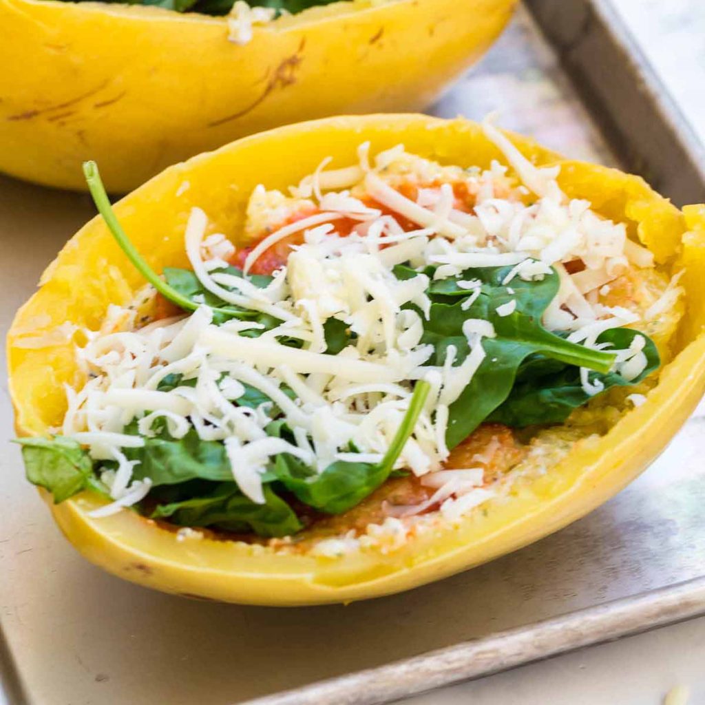 baking sheet with uncooked spaghetti squash halves filled with cottage cheese and greek yogurt mixture, then topped with tomato sauce, spinach and cheese.