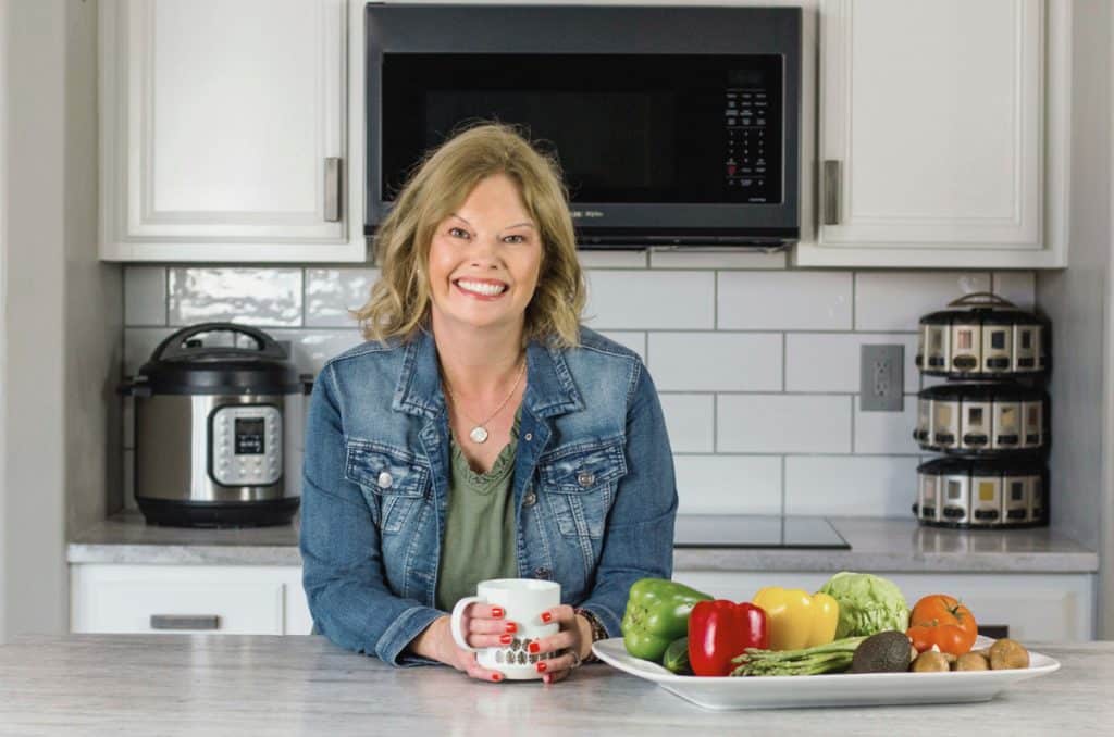 Becca in an olive green dress with a denim jacket, leaning over a white countertop and holding onto a coffee mug