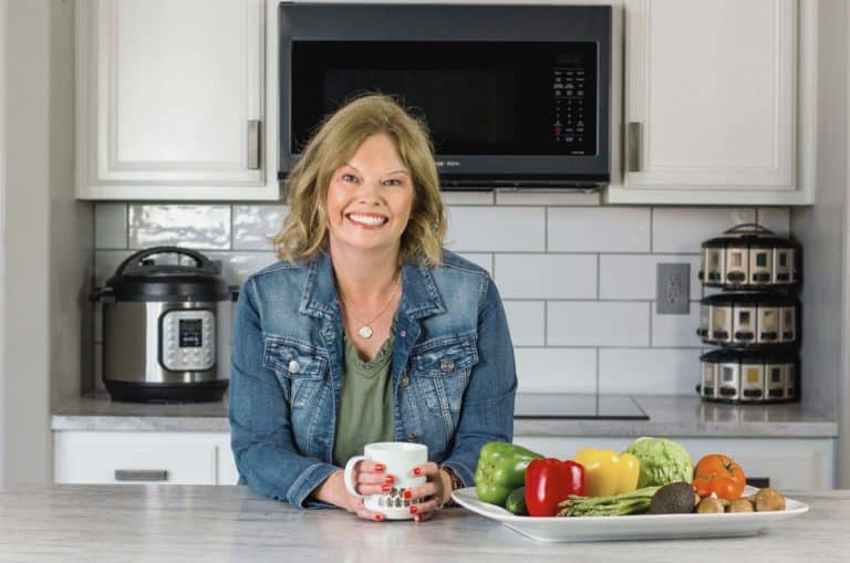 Becca in an olive green dress with a denim jacket, leaning over a white countertop and holding onto a coffee mug