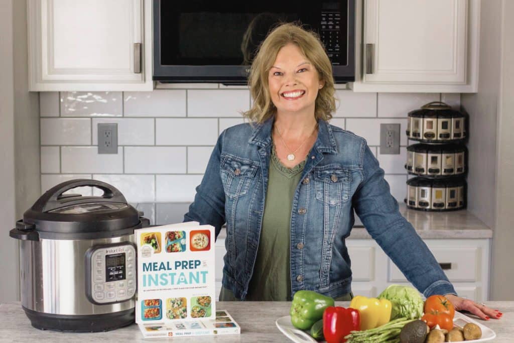 Becca is wearing an olive dress and a denim jacket. She has a white counter in front of her with an Instant pot, stack of books, and a tray of fresh veggies