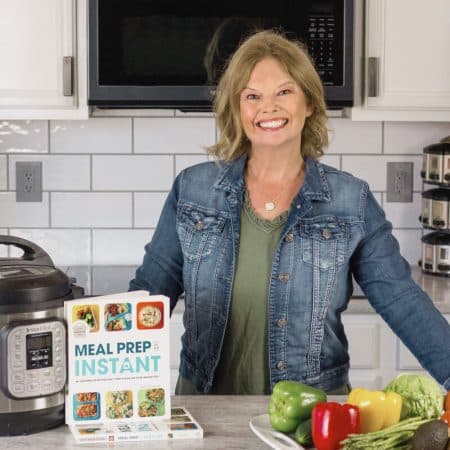 Becca is wearing an olive dress and a denim jacket. She has a white counter in front of her with an Instant pot, stack of books, and a tray of fresh veggies