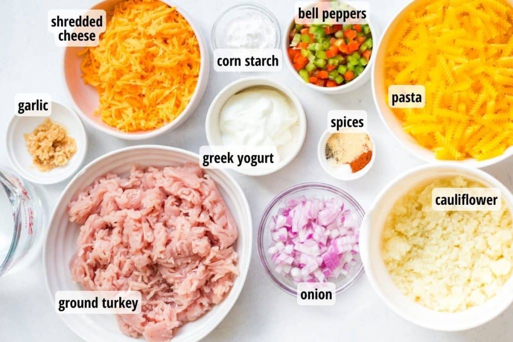 labeled ingredients needed to make philly cheesesteak pasta. Ingredients are in white and glass bowls on a white countertop.